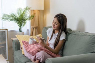 Cep telefonu kullanan gülümseyen genç bir kadın, akıllı telefondan keyif alan mutlu bir insan, evdeki kanepede rahat bir şekilde cep telefonuyla, elinde fincanla..