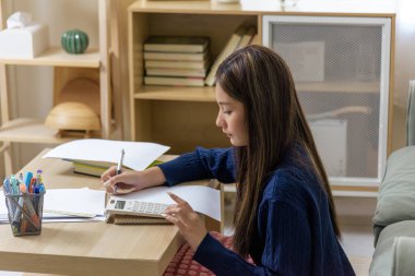 Öğrenci kız ödev yapıyor, hesap makinesi kullanarak çalışmak için notlar yazıyor, okul masasında sınava hazırlanıyor, genç Tayland 'lı kadın odaklanmış ifadeyle ciddi öğrenme odağı gösteriyor.