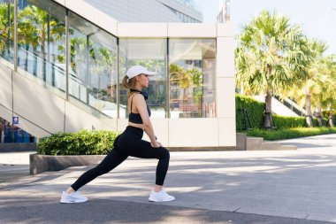 Spor kadını dışarıda antrenman yapıyor, formda olan kişi streç egzersizler yapıyor, aktif giyim sporcusu dinamik bacak esnemesini tamamlıyor, modern binalar ve palmiye ağaçları yakınlarındaki kentsel ortamda esneklik sağlıyor..