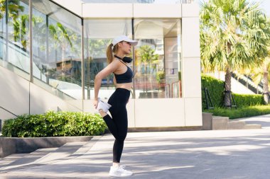 Spor kadını dışarıda egzersiz yapıyor, formda bireysel olarak modern mimarinin yanında güneşli bir günde sağlık ve genel spor performansı için ferahlatıcı esneme egzersizleri yapıyor..