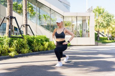 Formda bir kadın spor egzersizi yapıyor, kaslarını germek için hamle yapıyor, yeşil palmiye ağaçları ve modern binalarla kentsel ortamda sağlıklı bir vücut sergiliyor..