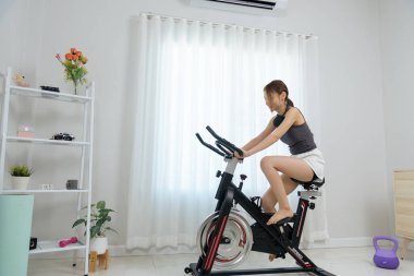 Evde genç bir kadın, mutlu bir insan fitness aktivitesinden, egzersiz bisikletinden, kulaklık takmaktan, sağlıklı yaşam tarzına odaklanmaktan, kişisel egzersiz hedeflerine ulaşmaktan, sağlık rutinini korumaktan hoşlanıyor..