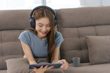 Evdeki genç mutlu kadın, tablette dijital aktiviteden zevk alan, kulaklıklı, kahverengi koltukta dinlenen, eğlenirken gülümseyen insanları temsil ediyor..