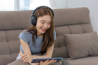 Genç mutlu kadın ev aktivitesi, insanlar rahatlama buluyor, kulaklık takıyor, tablet cihazından dijital içeriği izliyor, geniş bir gülümseme ile oturma odasının keyfini çıkarıyor..