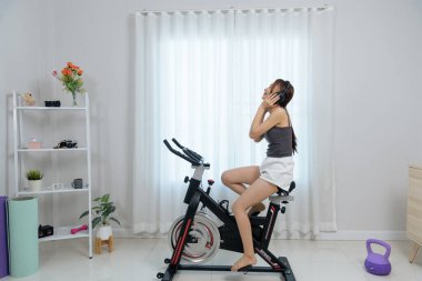 Genç bir kadın evde mutlu mesut spor yapıyor, bisiklet üzerinde egzersiz yapıyor, kulaklık takıyor, genel refah ve vücut gücü için sağlıklı ve enerjik iç egzersizlerin keyfini çıkarıyor..