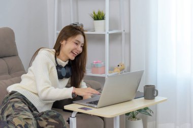 Genç kadın mutlu, insanlar için ev aktivitesi, bilgisayarla uzaktan kumanda, çevrimiçi etkileşim sırasında gülümseme, günlük kıyafet, kulaklık takmak.