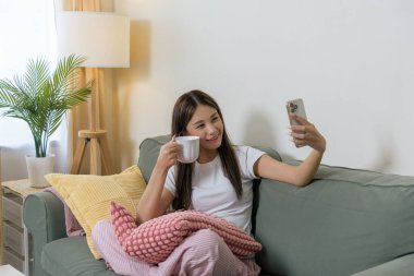 Cep telefonu kullanan mutlu bir kadın, mobil internet bağlantısından hoşlanan biri, ev koltuğunu gevşeten, sıcak bir içecek tutan, boş zamanı değerlendiren, rahat bir ortam hazırlayan biri..