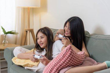 İki genç bayan arkadaş evde video oyunu oynuyor, gençler kontrolörlerle eğleniyor, gülümsüyor, gülüyor, rahat koltukta dinleniyor..