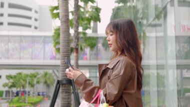Alışveriş çantalı mutlu genç kadın modern alışveriş merkezinin yanında duruyor, gülümseyen insan kıyafet alıyor, neşeli anıları yakalamak için telefonu tutuyor, açık havada eğlencenin tadını çıkarıyor..