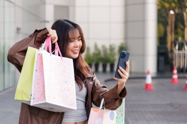 Asyalı kadın alışveriş yapmayı seviyor, alışveriş merkezinin yakınındaki başarılı mağaza gününden kalma moda çantalarını tutuyor, insanlar siyah cuma günü cep telefonu kullanarak yaşam tarzı deneyimlerini kucaklıyor..