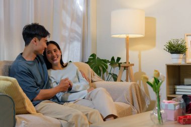 Evde aşık genç bir çift, kadın ve erkek romantik bir sevgililer günü geçiriyorlar, gülümsüyorlar ve rahat koltukta el ele tutuşuyorlar, hediyeler ve çiçeklerle samimi bir an paylaşıyorlar..