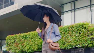 Genç iş kadını, elinde şemsiyeyle modern ofis seyahati için dışarıda dolaşmaktan mutlu. Elinde şık bir çanta, yemyeşil bitkilerle modern şehir ofis binasının önünden geçiyor..