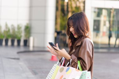 Asyalı bir kadın alışveriş yapmayı seviyor, insanlar için aktif bir yaşam tarzı, alışveriş merkezinin girişinde moda çantaları tutuyor, parlak bir şekilde gülümsüyor, siyah cuma günü dükkan olayından sonra dışarıda poz veriyor, cep telefonlarını kontrol ediyor..