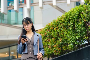 Genç kurumsal kadın şehir caddesinde yürüyor. Çalışmak için akıllı telefon kullanıyor. Yoğun şehir ortamında, modern bina ve arkasında yeşil yeşillikler var..