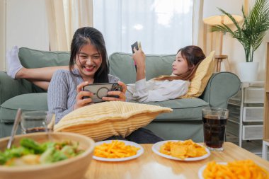 Genç iki kadın atıştırmalık yemekten hoşlanır, arkadaşı rahat oturma odasında dinlenir, bir kız gezici oyun oynar, diğeri masal kitabı okur, mutlu insanlar evde boş zaman aktivitelerini kucaklarlar..