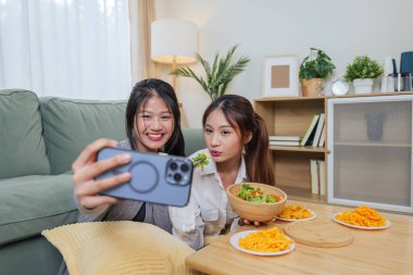 İki genç Asyalı kadın yemek yemekten hoşlanır, arkadaşlar akıllı telefonlarıyla selfie çekerler, rahat oturma odasında yemek yerler, kameraya gülümseyen mutlu insanlar, lezzetli sağlıklı salata ve lezzetli atıştırmalıklar yerler..