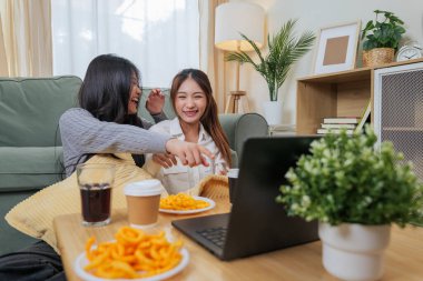 İki genç bayan arkadaş yemek yemekten, oturma odasında gülmekten, lezzetli atıştırmalıkları ve içecekleri paylaşmaktan, dizüstü bilgisayarları izlemekten, modern ev atmosferini rahatlatmaktan hoşlanırlar..
