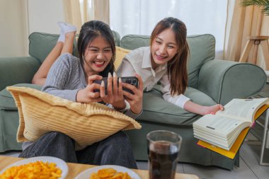 İki mutlu Asyalı genç bayan arkadaş lezzetli atıştırmalıklar yemekten, telefon eğlencesi izlemekten, birlikte gülmekten, rahat bir oturma odasında iç mekanda, modern ev hayatı tarzında vakit geçirmekten hoşlanırlar..