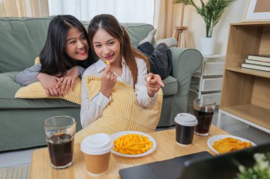 Genç kadınlar, iki arkadaş, rahat oturma odasında atıştırmalık yemekten, kahkahalar atmaktan ve neşeli etkileşimlerden hoşlanırlar. Bu arada dizüstü bilgisayardan medyanın içeriğini izleyerek, birlikte unutulmaz sıcak anlar yaratırlar..