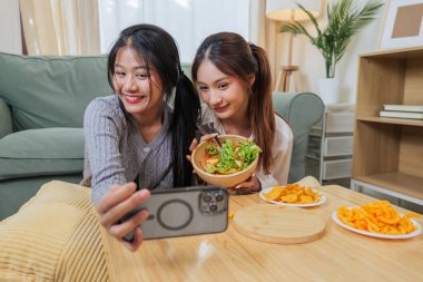 İki genç kadın sağlıklı salata yemekten, lezzetli atıştırmalıklar yemekten ve arkadaşlarıyla mutlu anları paylaşmaktan, modern oturma odasında dinlenmekten telefonla fotoğraf çekmekten, neşeli insanlarla kaynaşmaktan hoşlanırlar..