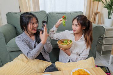 İki genç kadın sağlıklı yemek yemekten hoşlanıyor, arkadaşlar oturma odasında mutlu anları paylaşıyor, kız akıllı telefonlu taze salata fotoğrafları çekiyor, sosyal medya anılarını yakalıyor, evde rahat bir şekilde dinleniyor..