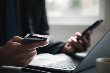 Bilgisayarın yanında kredi kartı ve akıllı telefon tutan bir elin yakın çekimi. Arka planda bir fincan kahve görünür.. .