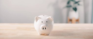 Beyaz seramik domuz kumbarası para biriktirmek ve iç mekanlarda finansal planlama için ahşap masada oturuyor, gelecekteki zenginlik için kişisel yatırımları ve ekonomik büyümeyi teşvik ediyor..