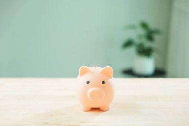 Para biriktirme hedefleri için pembe kumbara ahşap masaya oturur, kişisel finans için finansal yatırım konsepti, bulanık ev bitkisi ve minimalist ev içini gösteren arka plan.
