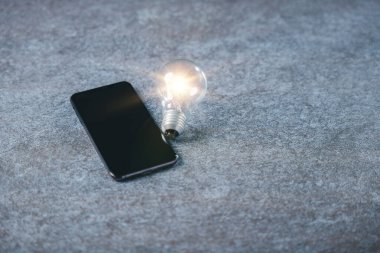 Parlayan ampul dijital pazarlama iş dünyasının yeniliklerine, pazar konsepti için yaratıcı bilgi geliştirmeye, gri dokulu arka planda akıllı telefonun yanı sıra stratejiye de yardımcı oluyor.