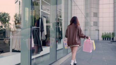 Bir sürü renkli çanta taşıyan, alışveriş merkezinden zevk alan, kıyafet alan, gülümseyen, modern şehir binasının önünden geçen genç ve mutlu bir kadın..