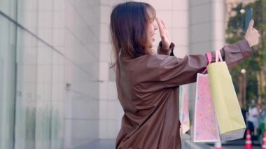 Alışveriş merkezinden alışveriş çantasıyla alışveriş yapan mutlu genç bir bayan. Modern binanın yakınındaki telefon görüşmesinde insanlar gülümsüyor ve el sallıyor..