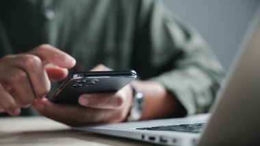 Arka planda dizüstü bilgisayarı olan birinin akıllı telefonunu tutarken yakın plan fotoğrafı. Görüntü akıllı telefonun kamera lensine odaklanıyor..