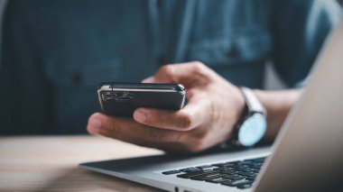 Arka planda dizüstü bilgisayarı olan bir kişinin eli akıllı telefonu tutuyor. Dizüstü bilgisayarın klavyesi odaklı.