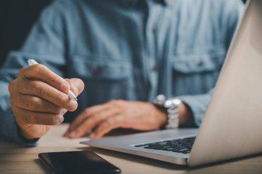 Ön planda dizüstü bilgisayarı ve akıllı telefonu olan bir stilus kalemi tutan elin yakın çekimi. Adam mavi bir gömlek giyiyor. İş, teknoloji veya üretkenlik temaları için mükemmel.