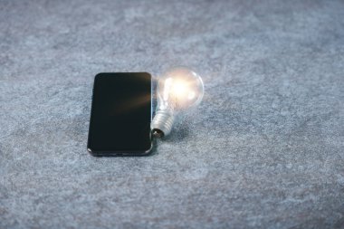 Dijital pazarlama fikri için parlayan ampul, gri taş yüzeydeki cep telefonu yanında modern iş yeniliği kavramı yaratıcı pazar gelişimi ve bilgisini gösteriyor.