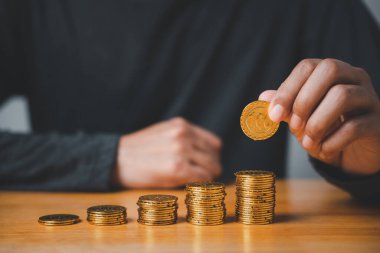 Bir el, altın bir madeni parayı büyüyen bir yığın altının üstüne koyar. Sikkeler ahşap bir masanın üzerinde..