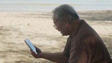 Yaşlı adam yaz tatilinde kumsalda mutlu bir şekilde seyahat etmeyi seviyor, akıllı telefon internet gezisi yapıyor, sıcak kumda, köpüklü okyanus suyunun yanında huzur içinde dinleniyor..