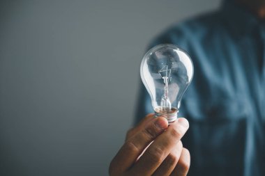 Ampulü tutan bir elin yakın çekimi. Ampul temiz ve iplik görünür durumda. Arka plan bulanık, ama mavi bir gömlek görülebilir..