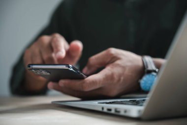 Arka planda dizüstü bilgisayarı olan bir masada otururken akıllı telefon kullanan birinin ellerini kapat..