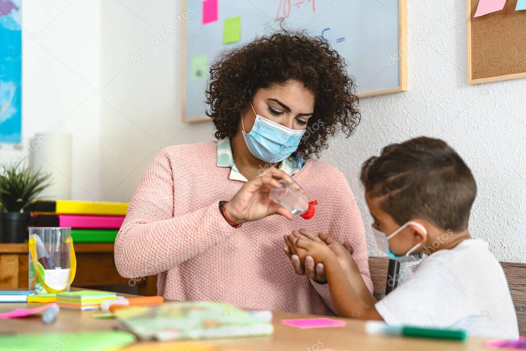 Teacher cleaning hands to stundet children with sanitizer gel while wearing face mask in preschool classroom during corona virus pandemic - Healthcare and education concept