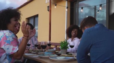 Aperatif hazırlayan ve teras partisinde birlikte kırmızı şarap içen çok kültürlü genç insanlar.