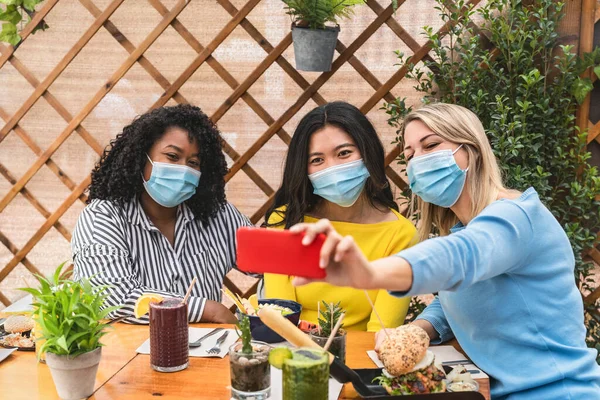 Multiracial friends wearing face mask while taking selfie with mobile smartphone cam in coffee brunch restaurant during corona virus outbreak