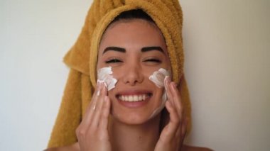 Gülümseyen genç bir kadın yüzüne krem sürüyor mutlu bir kız evde cilt bakımına gidiyor sağlıklı güzellik, temiz muamele ve gençlerin yaşam tarzı kavramı.