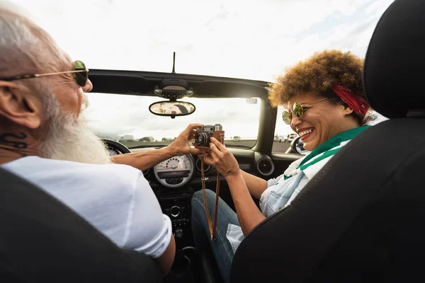 ロードトリップ中に新しいコンバーチブル車に乗っている間に古いヴィンテージカメラで写真を撮る楽しみを持って幸せなシニアカップル休暇 — ストック写真