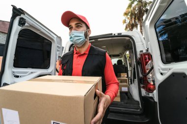Corona virüsünün yayılmasını önlemek için yüz maskesi takan karton kutu taşıyan genç teslimatçı Corona virüsü salgını sırasında hızlı bir şekilde yayılıyor.