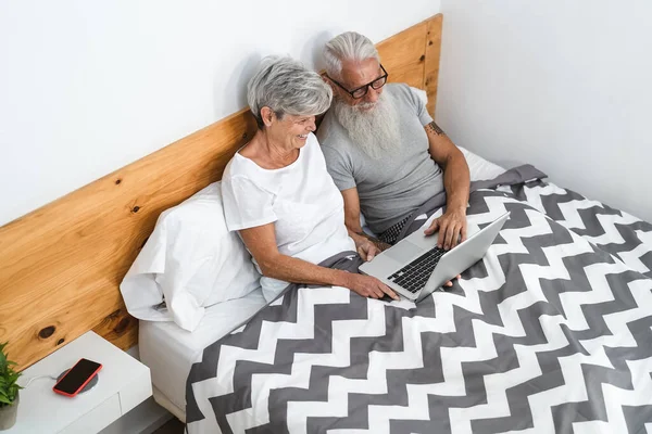 Happy senior couple having fun watching computer while lying in bed below blankets - Technology and elderly people lifestyle concept