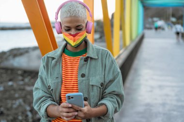 Genç eşcinsel kadın LGBT yüz maskesi takıyor ve kablosuz kulaklıkla müzik dinlerken cep telefonu kullanıyor - cinsiyet eşitliği ve teknoloji kavramı