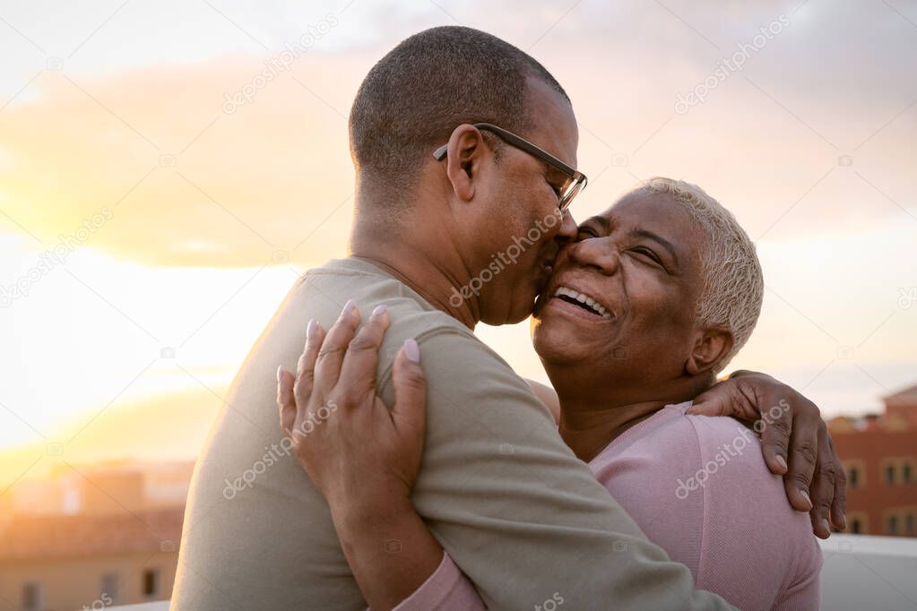 Happy Latin senior couple having romantic moment embracing on rooftop during sunset time - Elderly people love concept