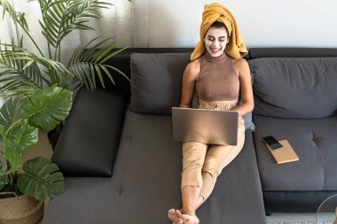 Evde cilt bakımı yaparken dizüstü bilgisayar kullanan genç bir kadın.