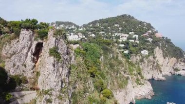 Aşağıdaki derin mavi deniz ile İtalya 'nın Capri Adası' nın dramatik kayalık kayalıklarını ve yemyeşil bitki örtüsünü bir hava manzarası yakalar. Binalar yamaçtaki ağaçların arasına kurulmuş..